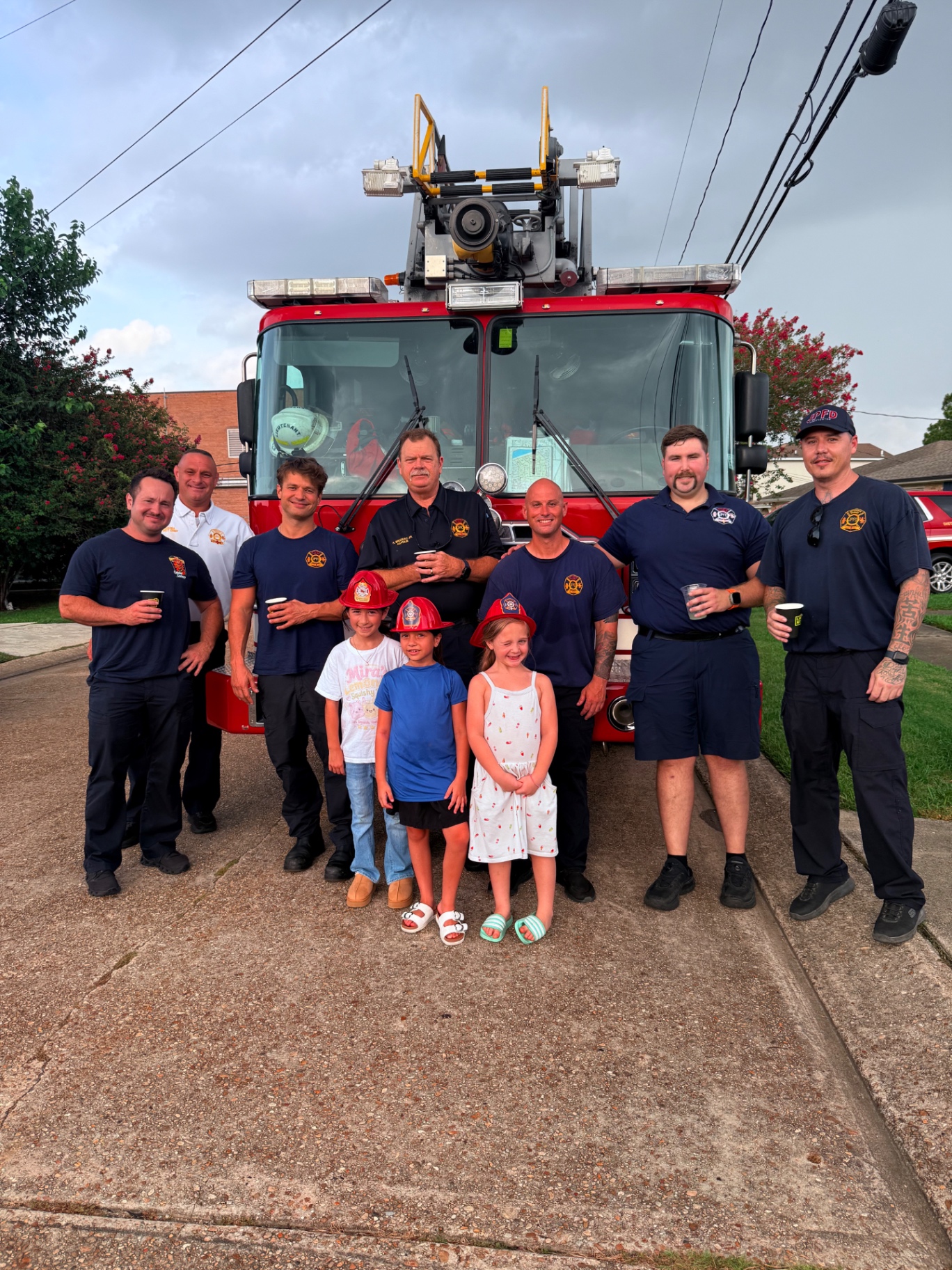 Seven Jefferson Parish firefighters in navy JPFD uniforms standing in front of a red ladder fire truck holding cups of Mira's fresh-squeezed lemonade, with Mira in her Mira's Lemonade Squishy t-shirt and two friends in red fire helmets smiling in front of them at the kid-run lemonade stand in Metairie, Louisiana