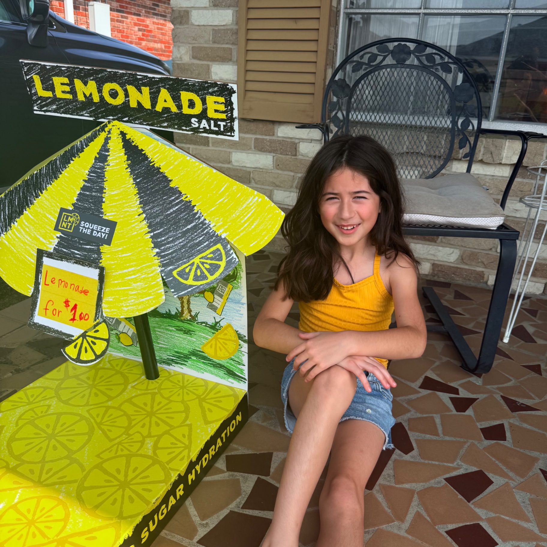 Young kid entrepreneur Mira smiling proudly next to her homemade yellow cardboard lemonade stand sign reading Lemonade $1.00 on the front porch β the very first Mira's Lemonade Squishy pop-up stand