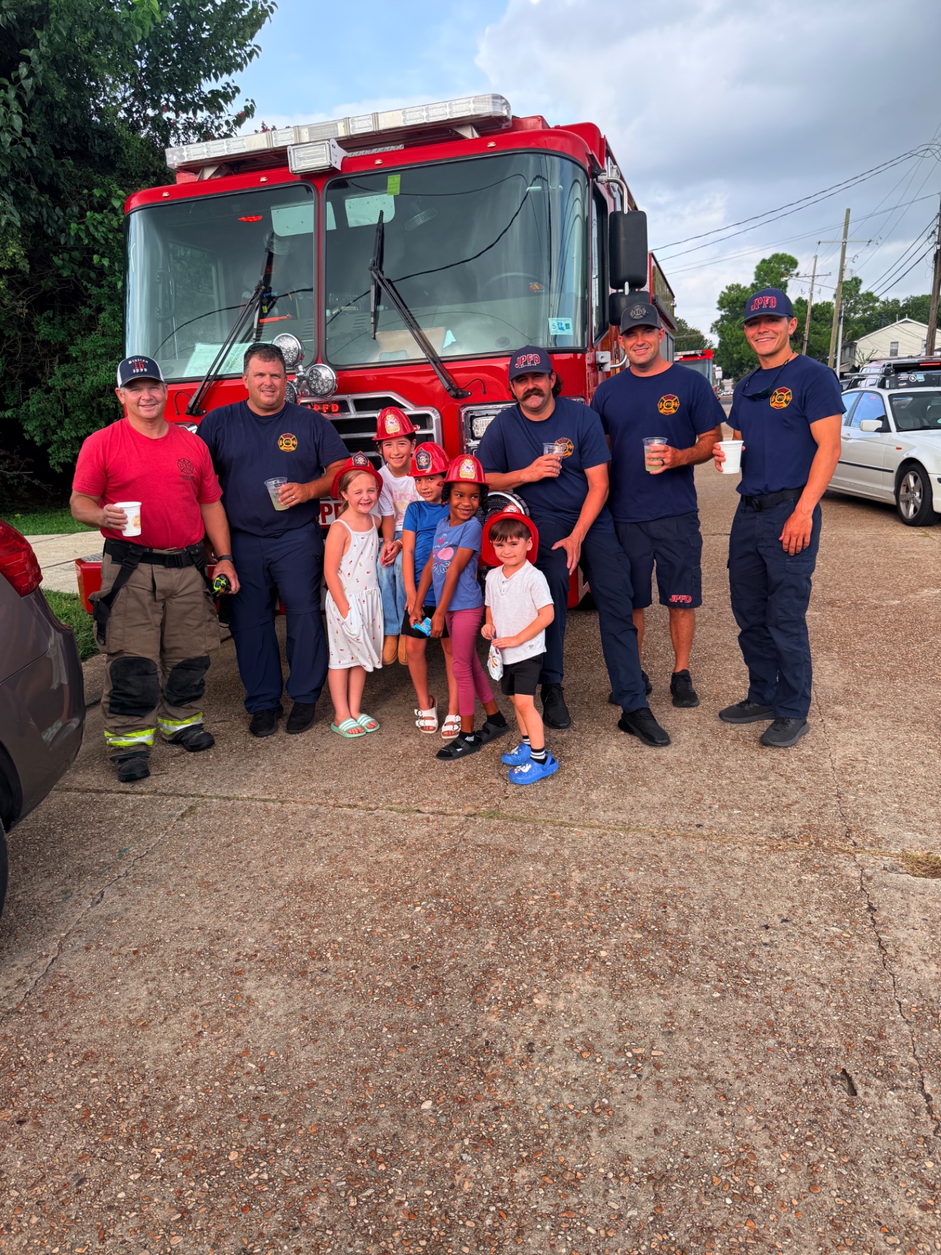 Five Jefferson Parish firefighters holding cups of lemonade beside a red JPFD fire engine while six kids in red plastic fire helmets, including Mira in her Mira's Lemonade Squishy shirt, pose in front at the Metairie lemonade stand and squishy rating table