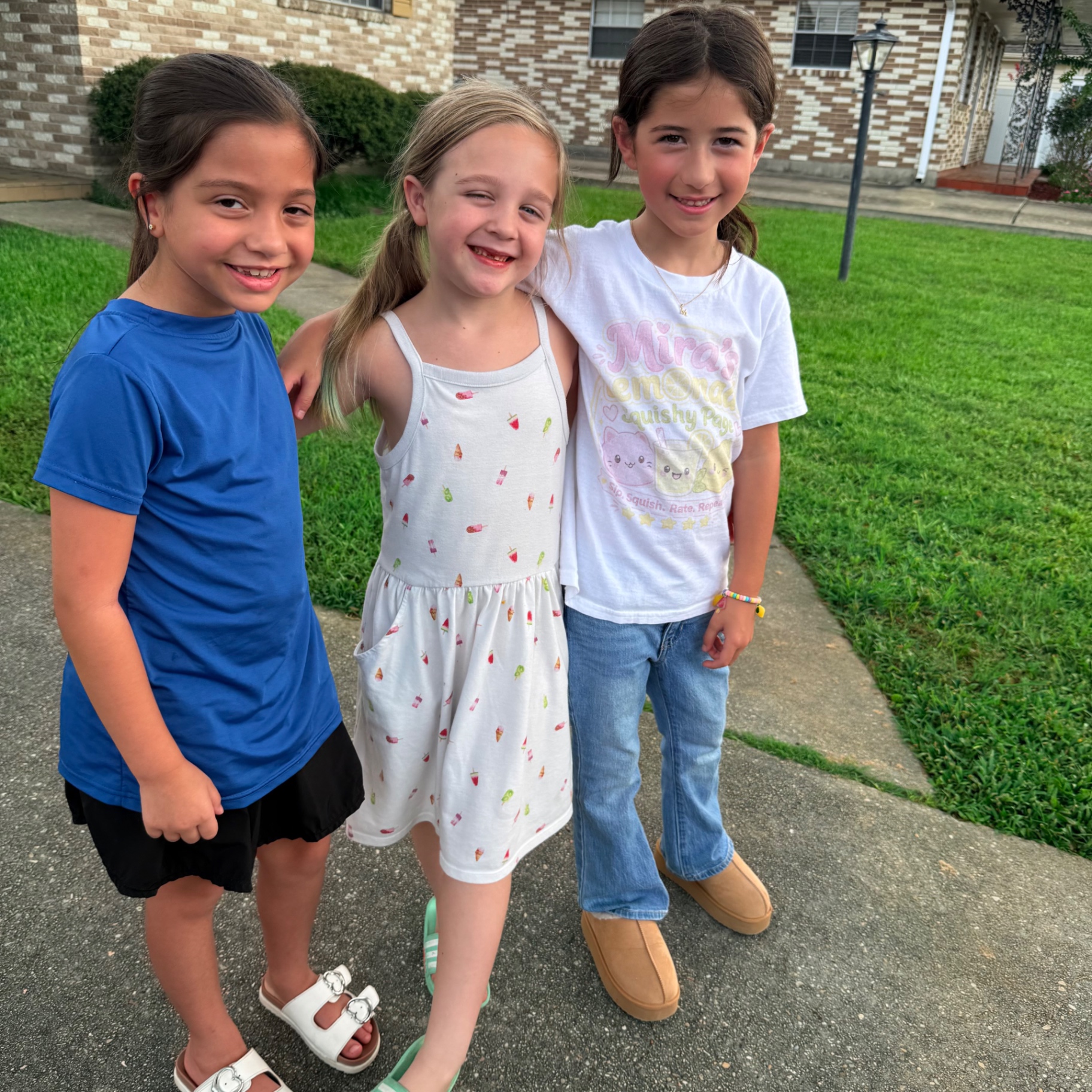 Mira in her white Mira's Lemonade Squishy t-shirt with the pink cat squishy and lemonade cup logo, standing arm-in-arm on the sidewalk with two friends after running the Metairie lemonade stand on firefighter visit day