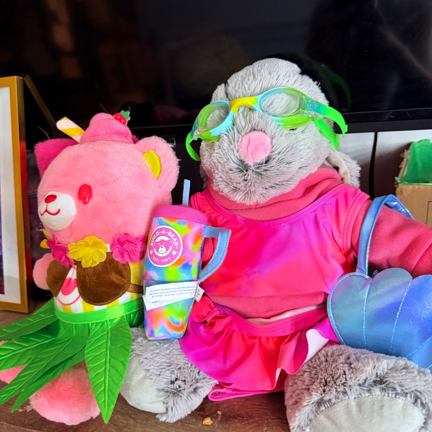 A soft pink teddy bear plushie sitting beside a gray bunny plushie wearing pink swim goggles, a pink dress, and holding a tie-dye Build-A-Bear cup β part of Mira's plushie and squishy collection
